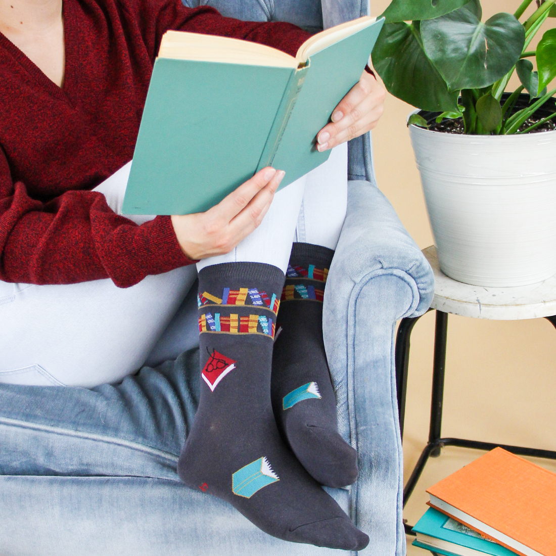 Photo of person curled up in the same blue armchair reading a green book and wearing dark grey socks with a pattern of books on them.