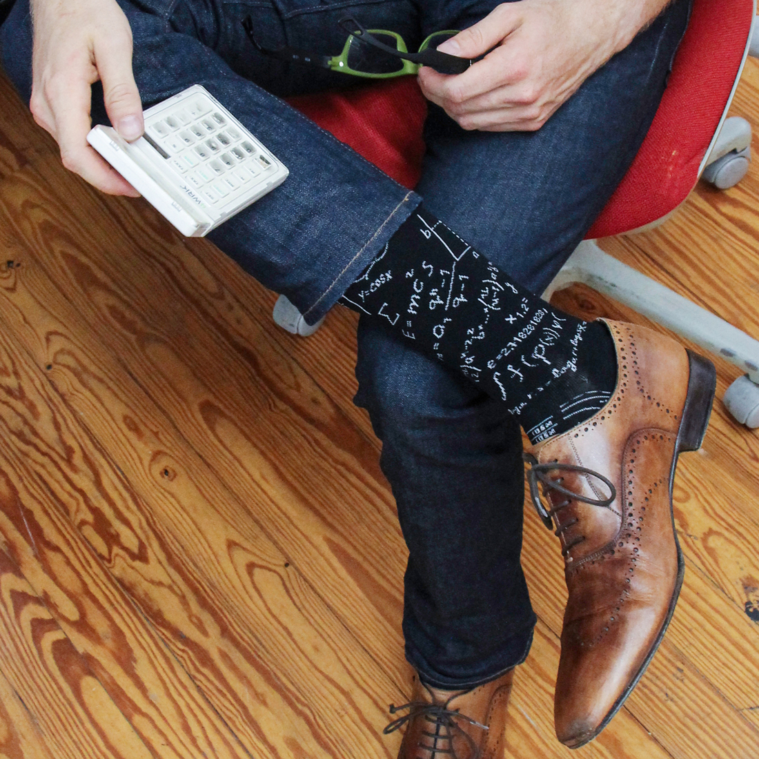 Photo of a man sitting in a red chair holding a calculator and green reading glasses with black socks that have random math equations scattered on them in white.