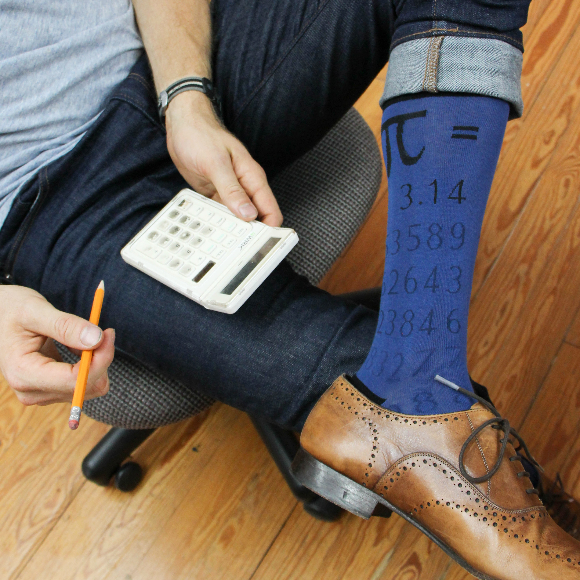 Photo of man sitting in chair holding a calculator and pencil, wearing jeans and dark blue socks that show the value of pi.