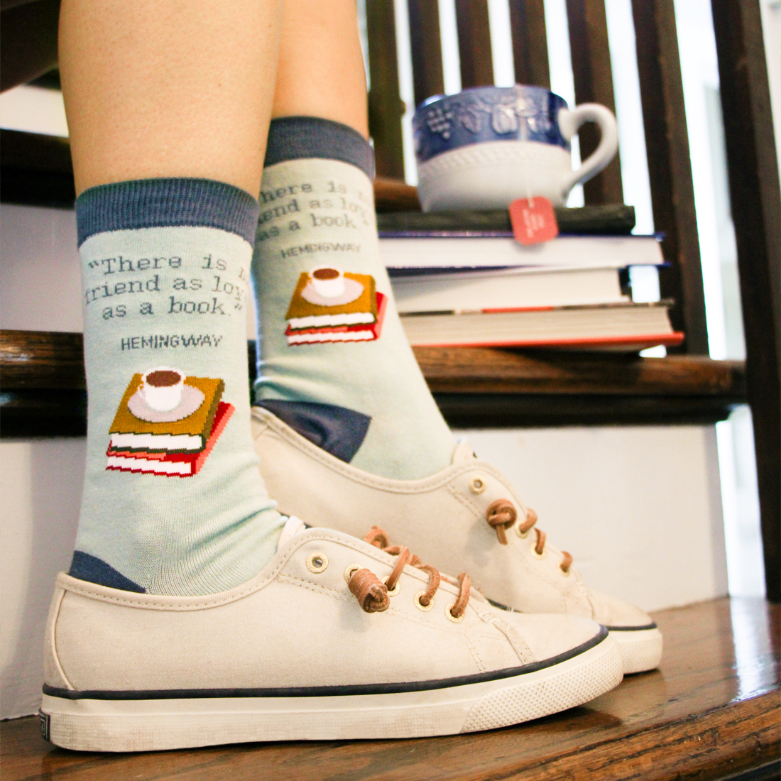Photo of feet wearing light blue socks that say, 'There is no friend as loyal as a book. Hemingway.' in white tennis shoes, on a wooden staircase with a stack of books and cup of tea.