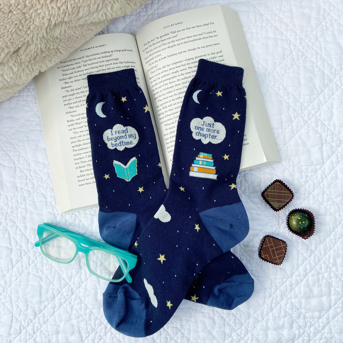 Photo of dark navy blue socks with a starry night and book design on them, laying on an open paperback book with tael reading glasses and some chocolate candies.