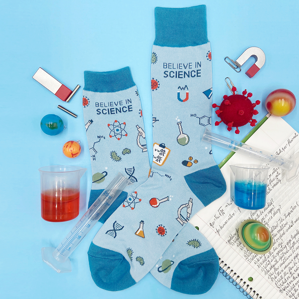 Photo of blue socks that have designs of science things like molecules and beakers and magnets that say, 'BELIEVE IN SCIENCE,' on a blue background with a notebook of chemistry notes, magnets, fake planet balls, and beakers filled with jello.