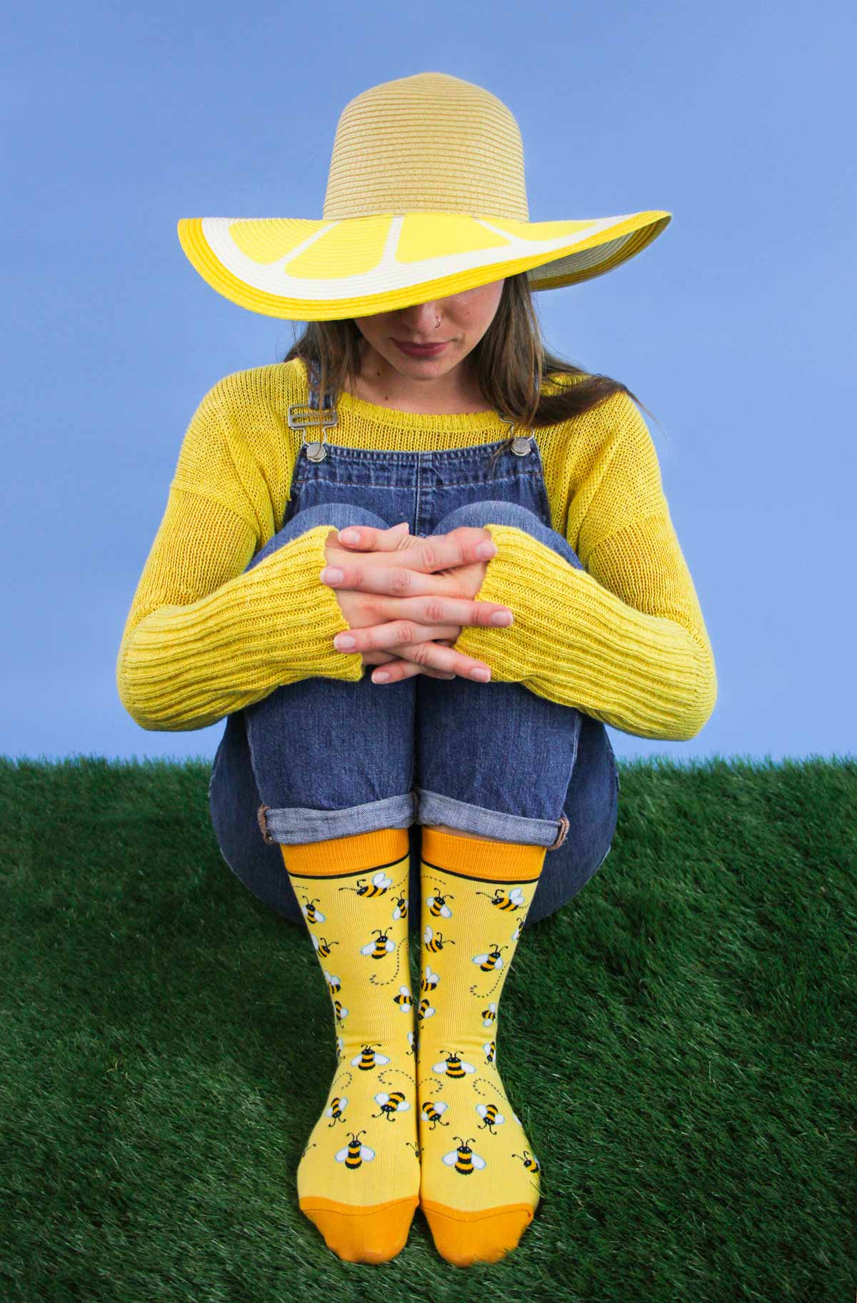 Woman in blue overalls wearing a yellow sweater, yellow sunhat, and bright yellow socks with bumblebee designs on them sitting on grass and a bright blue background.