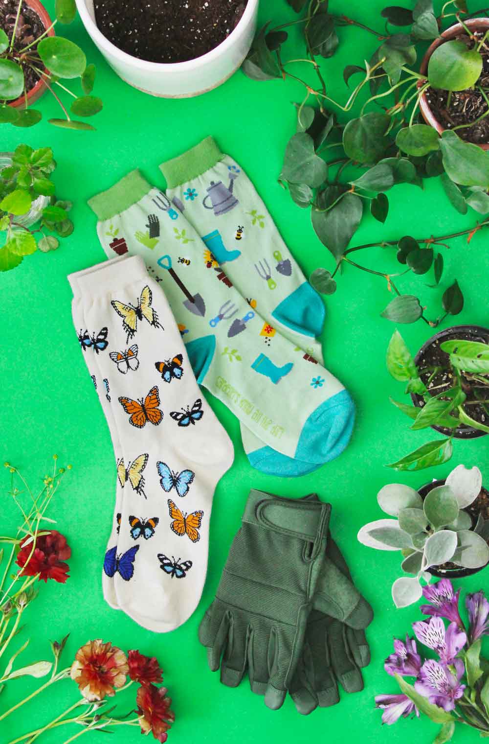 Butterflies and gardening socks laying on a green background surrounded by plants, flowers, and gardening gloves.