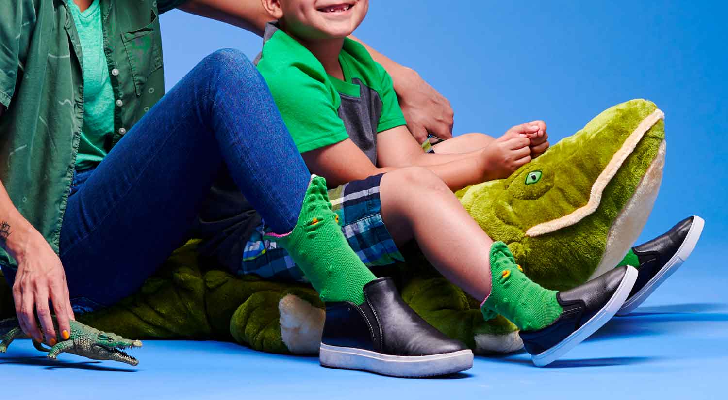 A woman and boy sitting on a giant plush stuffed alligator while wearing green 3D Alligator socks on a blue background.
