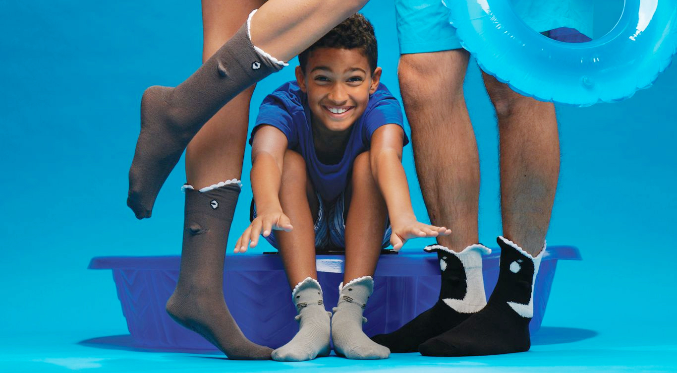 A cute little boy wearing Great White 3D socks jumping out from a blue kiddie pool, in between a woman wearing grey Shark Bite 3D socks and a man wearing 3D Killer Whale Orca socks holding a turquoise float tube.