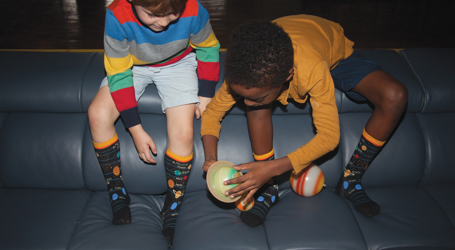 Two little boys wearing bright, fun clothes and matching kids planets socks, playing with miniature planet balls on a couch.