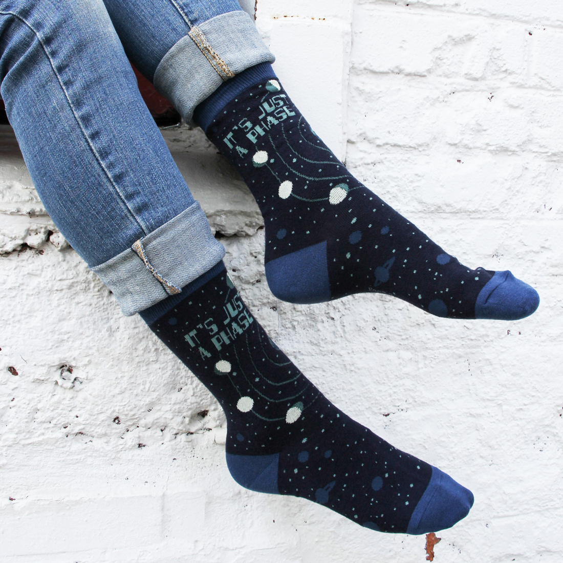 Woman in jeans wearing dark blue 'just a phase' moon phases socks sitting in front of a white brick wall.