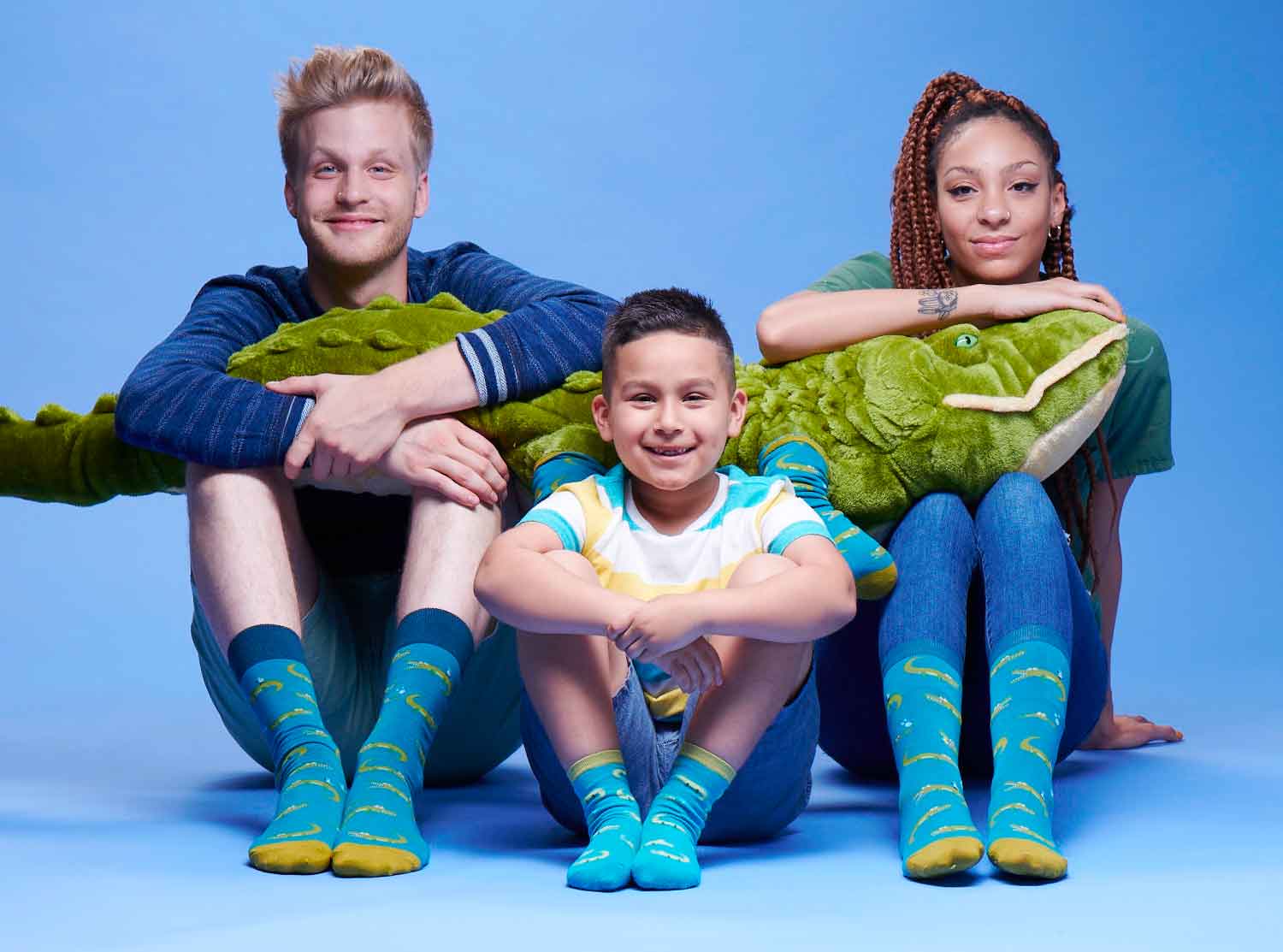 A man, kid, and woman sitting down looking at the camera wearing blue socks with alligators on them, holding a giant stuffed, plush alligator on a blue background.